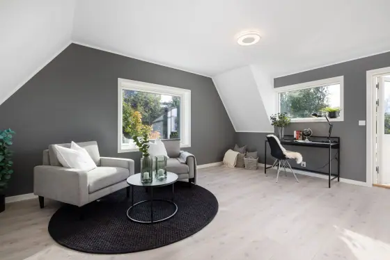 Loftstue under skråtak med sittegruppe, arbeidsplass og vinduer på to sider.