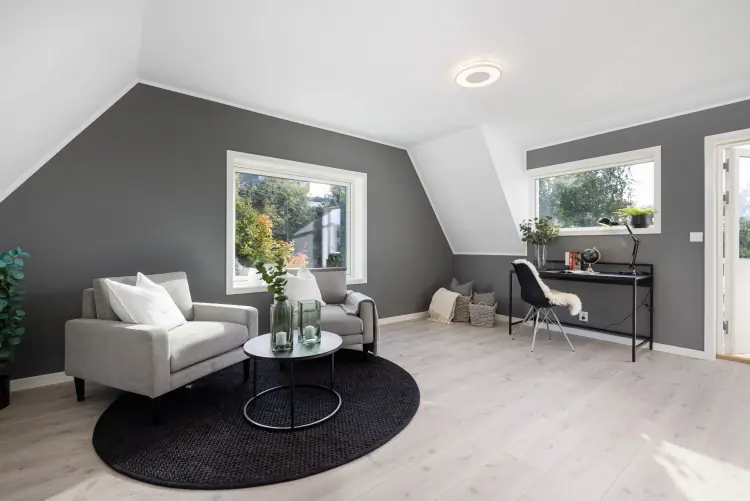 Loftstue under skråtak med sittegruppe, arbeidsplass og vinduer på to sider.